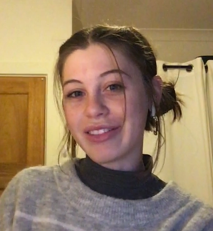 Maisie is smiling at the camera tilting their head. Their hair is tied up in two low buns. They are wearing a grey jumper.