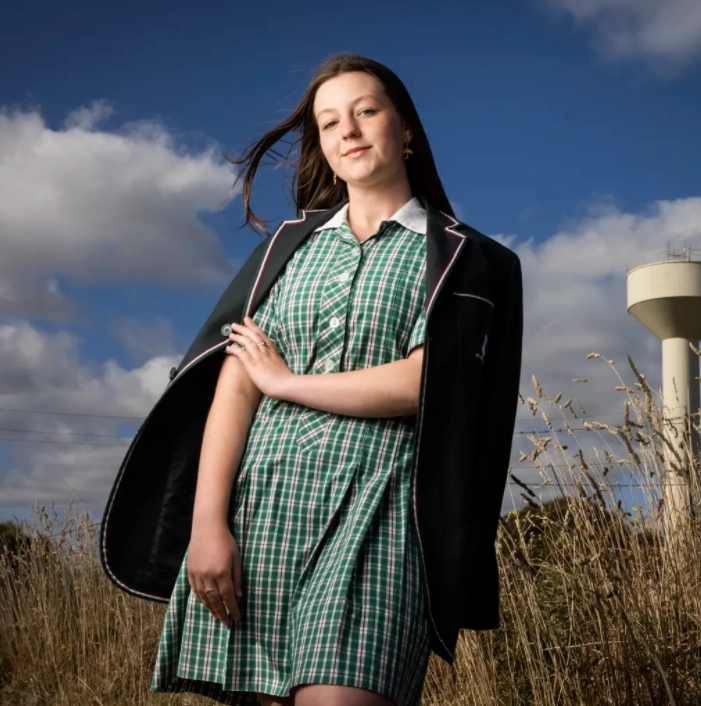 A school aged girl posing outside looking down onto camera, in a school dress and blazer with her hair swept up by the wind, posing confidently.