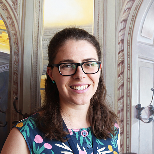 Benita, a woman with shoulder-length brown hair and glasses wearing a colourful dress standing in front of a painting of arches.