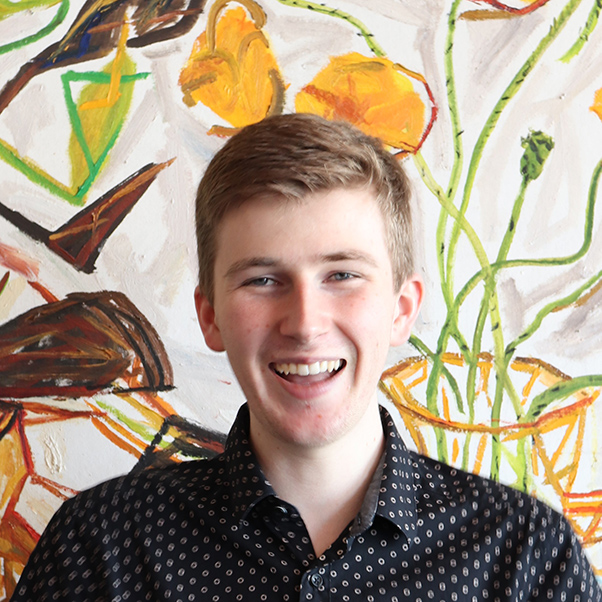 David, a young man with short, light brown hair in a patterned shirt standing in front of a painting.