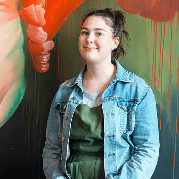Emily, a young woman with pulled back brown hair wearing a green blouse and denim jacket stands in front of a painting.