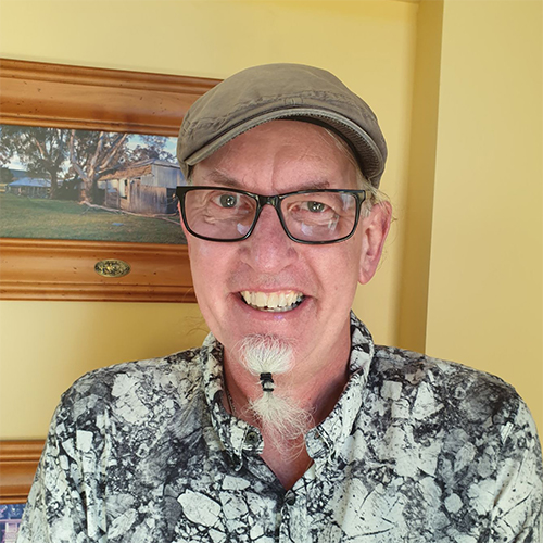 Ian, A man wearing a hat and glasses with a white beard in a ponytail stands in front of a wall.