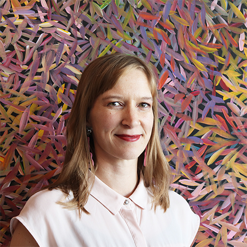 Nina, a woman with shoulder-length light brown hair and pink guitair earrings stands in front of a colourful painting.